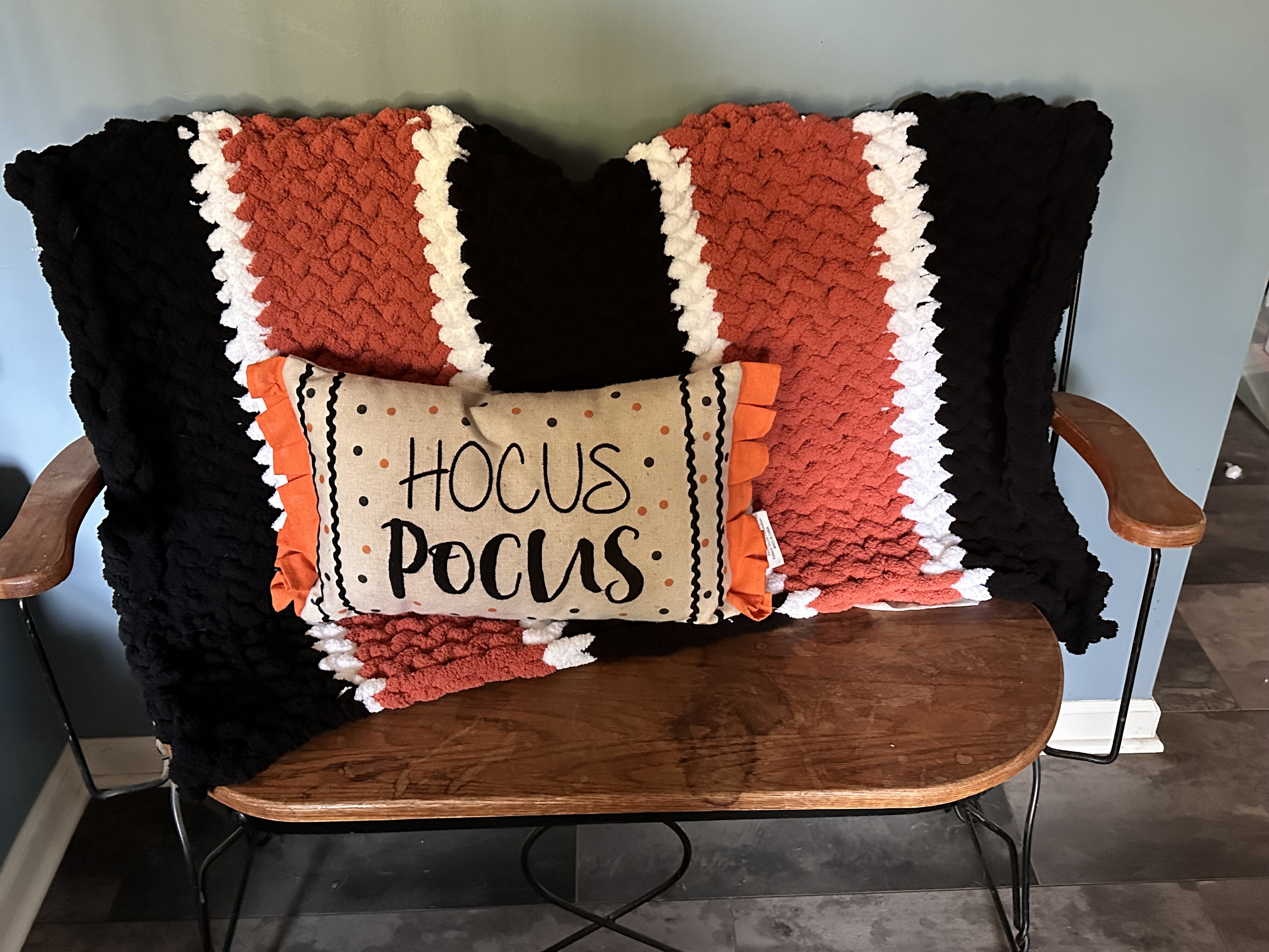 Black, burnt orange, and cream chunky knit blanket on a wooden bench with a Hocus Pocus pillow